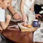A child cooks with his mom, helping to crack eggs at a table showing cooking with kids